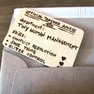 Può includere: Un biglietto di legno con il testo "OFFICIAL TEACHER ACCESS" e "TINY HUMAN MANAGEMENT" è parzialmente inserito in una busta grigia. Il biglietto elenca le competenze: "CONFLICT RESOLUTION", "SHOE TYING" e "GLITTER CONTROL".