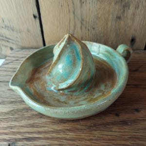 May include: Handmade ceramic citrus juicer in shades of green, brown, and turquoise. The juicer features a conical reamer in the center, a spout, and a handle. The ceramic piece is glazed and sits on a wooden surface.