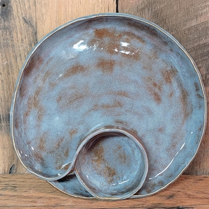 May include: A handmade ceramic serving dish with a unique design. The dish features a large, round plate with a smaller, circular bowl attached. The glaze is a blend of blue and brown, creating a marbled effect. The rim has a gold accent.