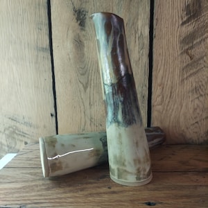 May include: Two handmade ceramic pitchers with a rustic design. The tall pitcher is brown, white, and tan, and the shorter pitcher is tan and white. The pitchers are on a wooden surface.
