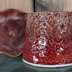 May include: A ceramic candle holder and matching dish. The candle holder is a deep red with a white base and star-shaped cutouts. The dish is a similar color with a scalloped edge.