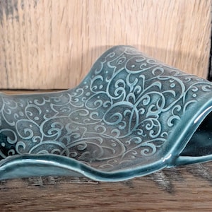 May include: A teal ceramic soap dish with a scrolled design. The dish has a curved, wave-like shape with a raised edge. The surface is glossy and the design is a lighter shade of teal.
