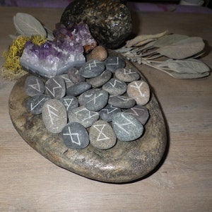 May include: A set of grey stones with white runes carved into them, arranged in a circle on a grey stone plate. The stones are surrounded by purple amethyst crystals and dried sage.
