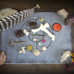 May include: A blue denim cloth with a circle drawn on it. The circle is filled with various natural objects, including bones, feathers, stones, and a small wooden disc with a red center.