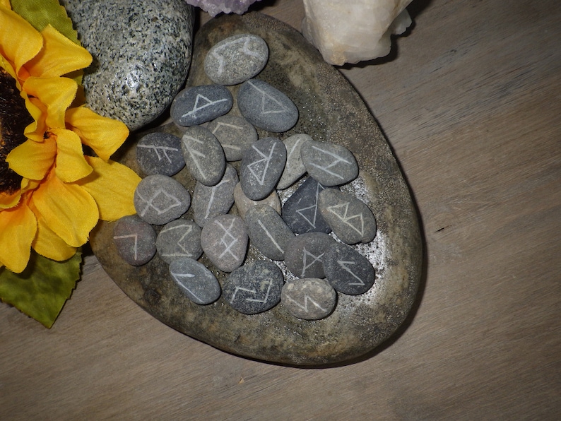 May include: A set of gray rune stones with white markings, arranged in a circle on a gray stone dish. The stones are surrounded by a sunflower.