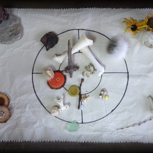 May include: A white cloth with a black circle drawn on it. The circle is divided into four sections with a bone in each section. There are other objects on the cloth, including a small piece of fur, a small piece of wood, a small piece of glass, and a small piece of metal.