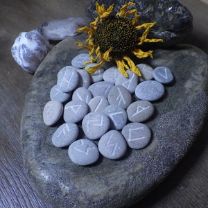 May include: A set of grey stones with white runes carved into them, arranged on a grey stone plate. A dried sunflower with yellow petals is on top of the stones.