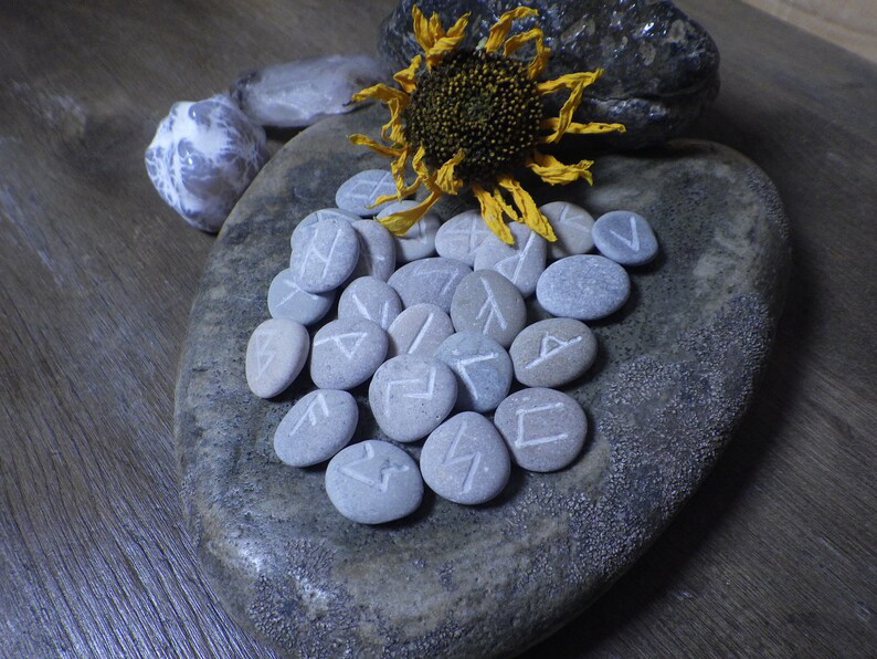 May include: A set of grey stones with white runes carved into them, arranged on a grey stone plate. A dried sunflower with yellow petals is on top of the stones.
