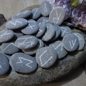 May include: A set of grey stones with white runes painted on them. The stones are arranged on a grey rock with a rough texture.