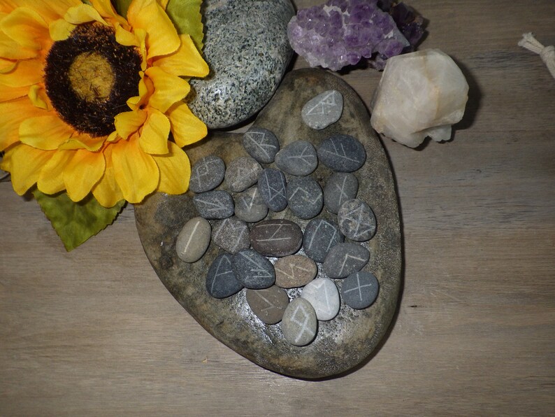 May include: A heart-shaped stone with gray and brown markings holds a collection of gray stones with white runes carved into them.