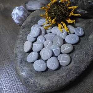 May include: A collection of gray stones with white runes painted on them, arranged on a gray stone surface. A dried sunflower with a yellow center is in the background.