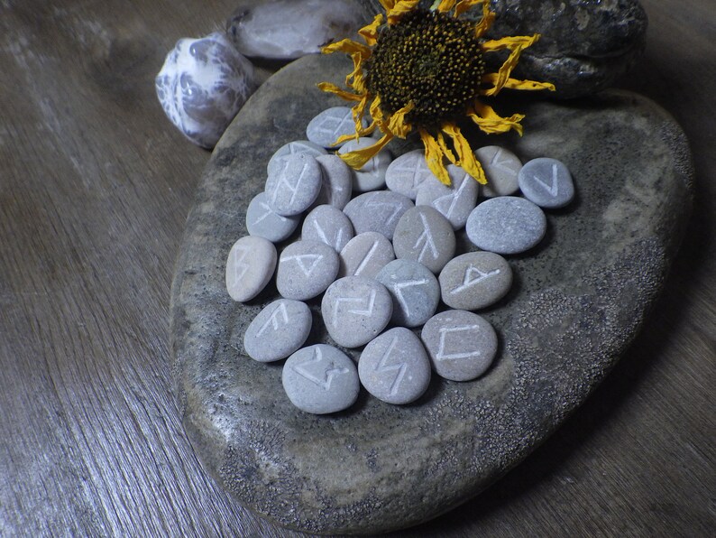 May include: A collection of gray stones with white runes painted on them, arranged on a gray stone surface. A dried sunflower with a yellow center is in the background.