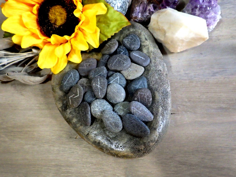 May include: A heart-shaped stone dish filled with gray and brown stones, each with a white rune carved into it. The dish is surrounded by a sunflower, a white crystal, and amethyst crystals.