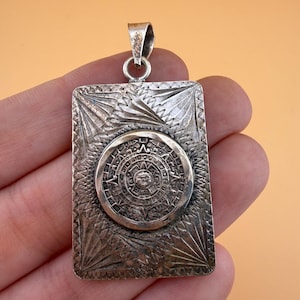 May include: A rectangular silver pendant featuring a central circular medallion with an embossed Aztec calendar design. The surrounding rectangular plate has a textured, sunburst-like pattern radiating from the center to the corners.