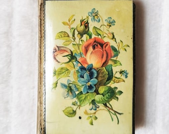 May include: A small, vintage book with a floral design on the cover. The cover features a bouquet of pink and red roses, blue flowers, and green leaves on a cream background. The book has a gold border.