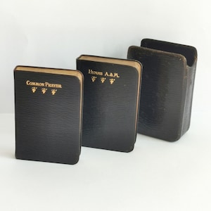 May include: Three small black leather-bound books with gold lettering. The books are titled "Common Prayer", "Hymns A & M", and are arranged from left to right.