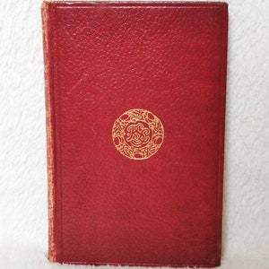 Pode incluir: Um livro encadernado em couro vermelho com um círculo em relevo dourado na capa. O círculo contém um desenho floral e as letras "PRS".