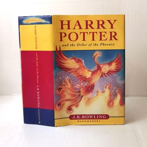 Puede incluir: Una cubierta de libro amarilla, roja y azul con el título "Harry Potter y la Orden del Fénix" y el nombre del autor "J.K. Rowling". La cubierta presenta un fénix en vuelo con llamas detrás.