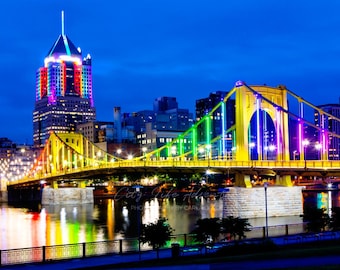 May include: Nighttime cityscape featuring a brightly lit bridge and skyscraper. The bridge is illuminated with colorful lights, reflecting in the water below. The sky is a deep blue, creating a vibrant contrast.