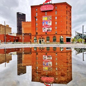 May include: A red brick building with a large Heinz ketchup bottle sign on top. The building is reflected in a puddle of water in the foreground. The building has a large sign that says "We Can Do It! Heinz History Center The Extraordinary Home of Pittsburgh".
