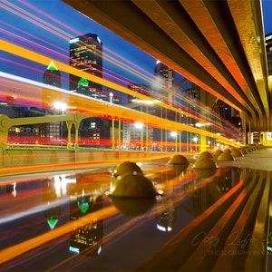 May include: A long exposure photograph of a bridge at night, with streaks of light from passing vehicles. The bridge has yellow support beams and a row of yellow bollards. City buildings are visible in the background, with a blue and orange sky.