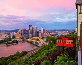 May include: A red vintage cable car with windows overlooking a cityscape at sunset. The city skyline features tall buildings and a yellow bridge. The cable car is on a track with a red railing.