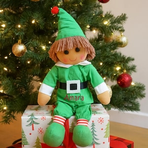 Peut inclure: Une poupée elfe verte avec une perruque en fil marron et un chapeau à pompon rouge est assise sur un coffret cadeau de Noël. La poupée porte une tenue verte avec une ceinture noire et le nom "Amara" est imprimé sur le devant. Le coffret est emballé dans du papier festif.