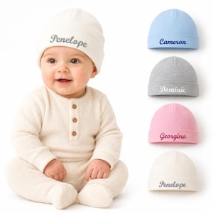 May include: A collection of five infant beanies in cream, light blue, grey, pink, and white. Each beanie features a folded brim and is embroidered with a unique name in cursive script, including Penelope, Cameron, Dominic, and Georgina.