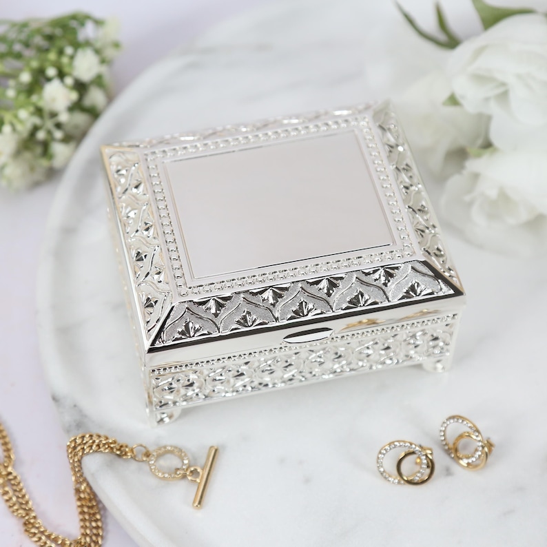 May include: A silver jewelry box with an ornate design. The box has a hinged lid and is decorated with a raised pattern. The box is sitting on a white surface.