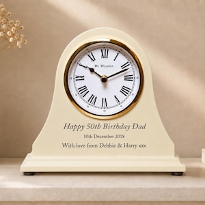 May include: A cream-colored mantel clock with a gold-toned bezel and a white face featuring black Roman numerals and the text "De Walden". The clock is engraved with "Happy 50th Birthday Dad", "10th December 2018", and "With love from Debbie & Harry xxx".