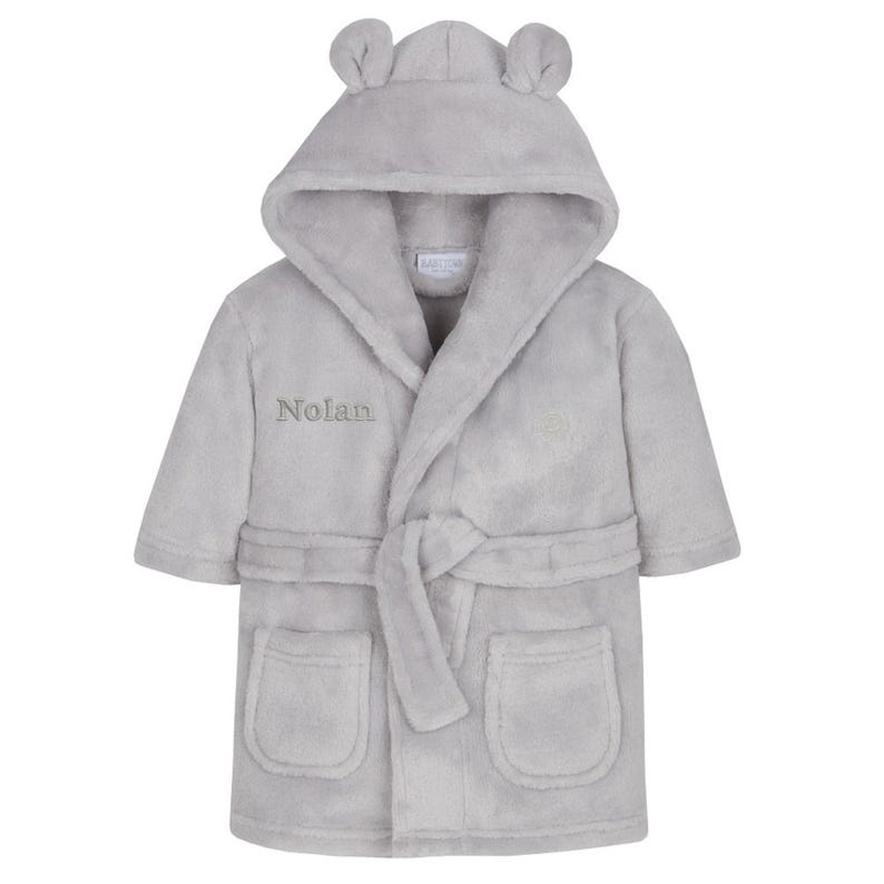 May include: A light gray plush hooded bathrobe with bear ears on the hood. The robe has a matching belt, two front pockets, and the name "Nolan" embroidered on the front. A cozy and comfortable children's garment.