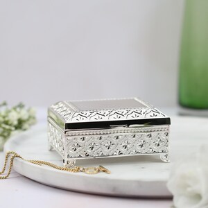 May include: A silver jewelry box with an ornate design. The box has a hinged lid and sits on four small feet.