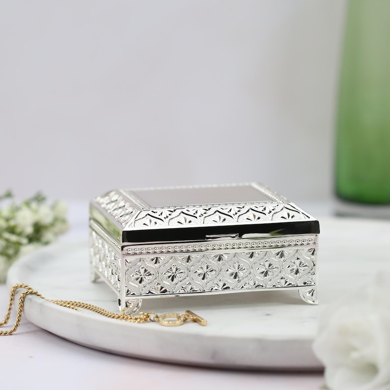 May include: A silver jewelry box with an ornate design. The box has a hinged lid and sits on four small feet.