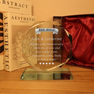 May include: A clear crystal wedding anniversary plaque with a geometric design. The plaque has the words "Wedding Anniversary" at the top, followed by names and a message. The base is rectangular. The plaque is displayed on a wooden surface.