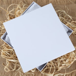 May include: A white cardboard box with a lid open, revealing a layer of white tissue paper. The box is sitting on a brown burlap surface with straw-like material around it.