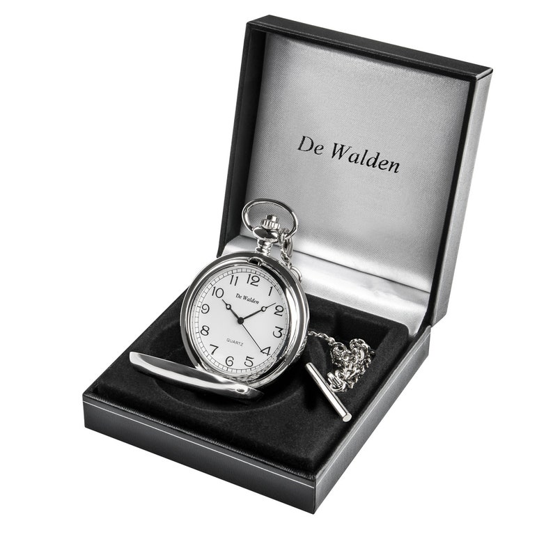 May include: A silver pocket watch with a white face and black numbers in a black velvet box. The watch has a chain and a fob. The box has the words "De Walden" printed on the inside lid.