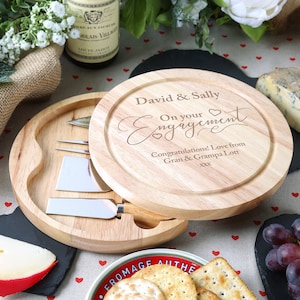 K&ouml;nnte beinhalten: Ein rundes, h&ouml;lzernes K&auml;seboard mit Deckel und einem Satz Messer im Inneren. Der Deckel ist mit dem Text "David & Sally On your Engagement Congratulations! Love from Gran & Grampa Lott xxx" graviert.