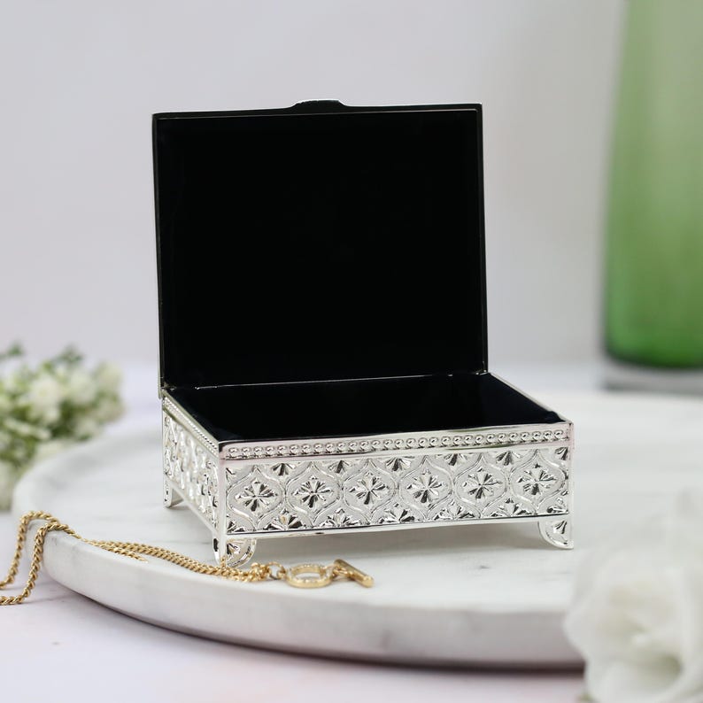 May include: A silver metal trinket box with an ornate design and a black velvet interior. The box is open and has a small gold chain resting on a white marble surface.