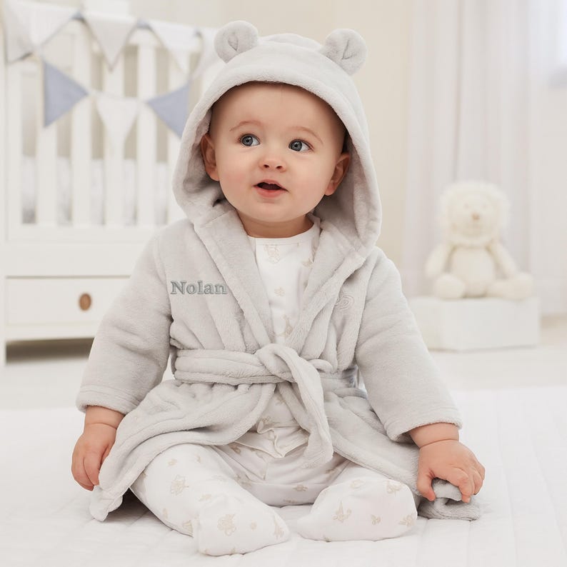 May include: A baby wearing a light gray hooded robe with bear ears and the name "Nolan" embroidered on the front. The robe is tied at the waist and the baby is wearing matching pajamas. A white stuffed animal is in the background.