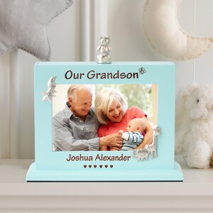 May include: A light blue photo frame with the words "Our Grandson" and "Joshua Alexander" in brown. The frame holds a photo of a baby with grandparents. A silver bear figurine sits on top, with decorative animals on the frame.