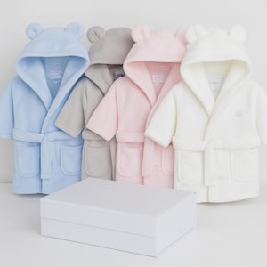 May include: Four plush baby bathrobes in pastel colors: light blue, gray, pink, and white. Each robe has a hood with bear ears, a matching belt, and pockets. A white gift box sits in front of the robes.
