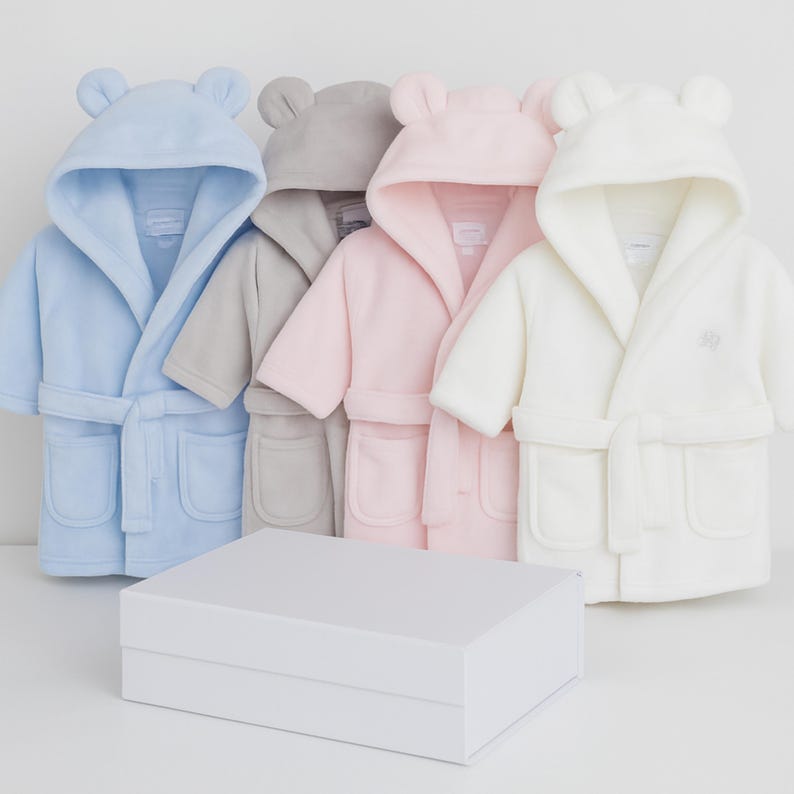 May include: Four plush baby bathrobes in pastel colors: light blue, gray, pink, and white. Each robe has a hood with bear ears, a matching belt, and pockets. A white gift box sits in front of the robes.