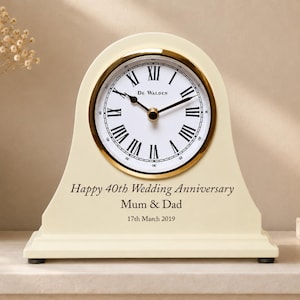 May include: A cream-colored mantel clock with a gold-toned bezel and a white face featuring black Roman numerals and the text "De Walden". The base is engraved with "Happy 40th Wedding Anniversary", "Mum & Dad", and "17th March 2019".