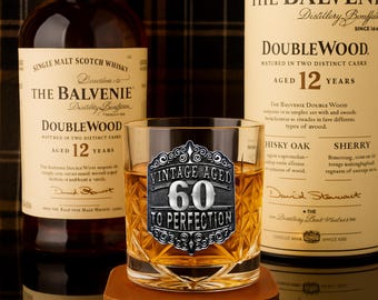 May include: A rocks glass with a silver "Vintage Aged 60 To Perfection" emblem, filled with amber liquid, sits on a brown coaster. Two bottles of The Balvenie Doublewood 12 Year Old Scotch Whisky are in the background.