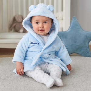 May include: A baby wearing a light blue hooded bathrobe with bear ears and the name "Harry" embroidered on the front. The baby is also wearing white patterned footed pajamas. A blue star-shaped pillow is in the background.