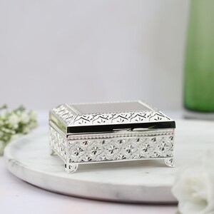 May include: A silver decorative trinket box with an ornate design. The box has a hinged lid and sits on four small feet.