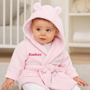 May include: A baby wearing a light pink hooded bathrobe with bear ears and the name "Ember" embroidered on the front. The baby is also wearing white and gray polka dot footed pajamas. The robe is tied at the waist.