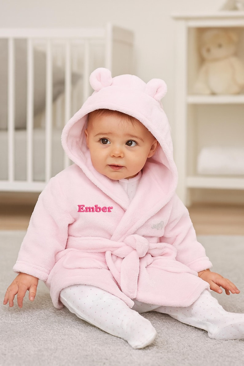 May include: A baby wearing a light pink hooded bathrobe with bear ears and the name "Ember" embroidered on the front. The baby is also wearing white and gray polka dot footed pajamas. The robe is tied at the waist.