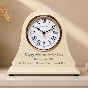 May include: A cream-coloured arched mantel clock with a gold-tone bezel and a white face featuring black Roman numerals and the text De Walden. The base is engraved with Happy 90th Birthday Dad, 17th December 2018, and With love from Dennis, Alisha, Fran & Dan xx.