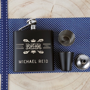 May include: Black stainless steel hip flask with a silver lid and funnel. The flask is engraved with the text "ESTD. 1984" and "MR" with a crown above the letters. The text "MICHAEL REID" is engraved below the logo. The flask is on a blue and white polka dot fabric.